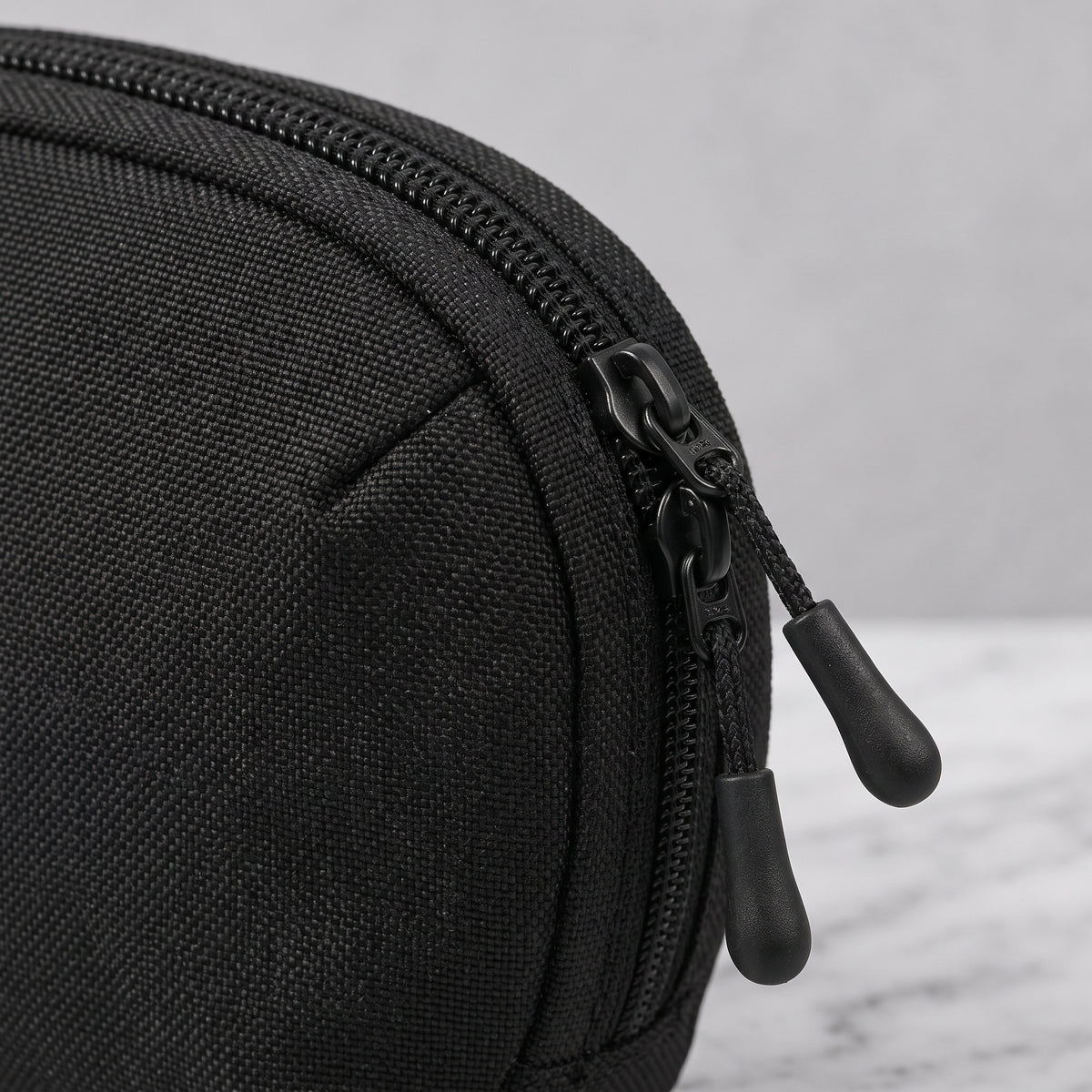 Nahaufnahme eines schwarzen Organizer Pouch aus robustem, strukturiertem Textilgewebe mit schwarzem Doppel-Reißverschluss und zwei Zipper-Anhängern, platziert ist die Aufbewahrungstasche auf hellem Untergrund in Marmoroptik.