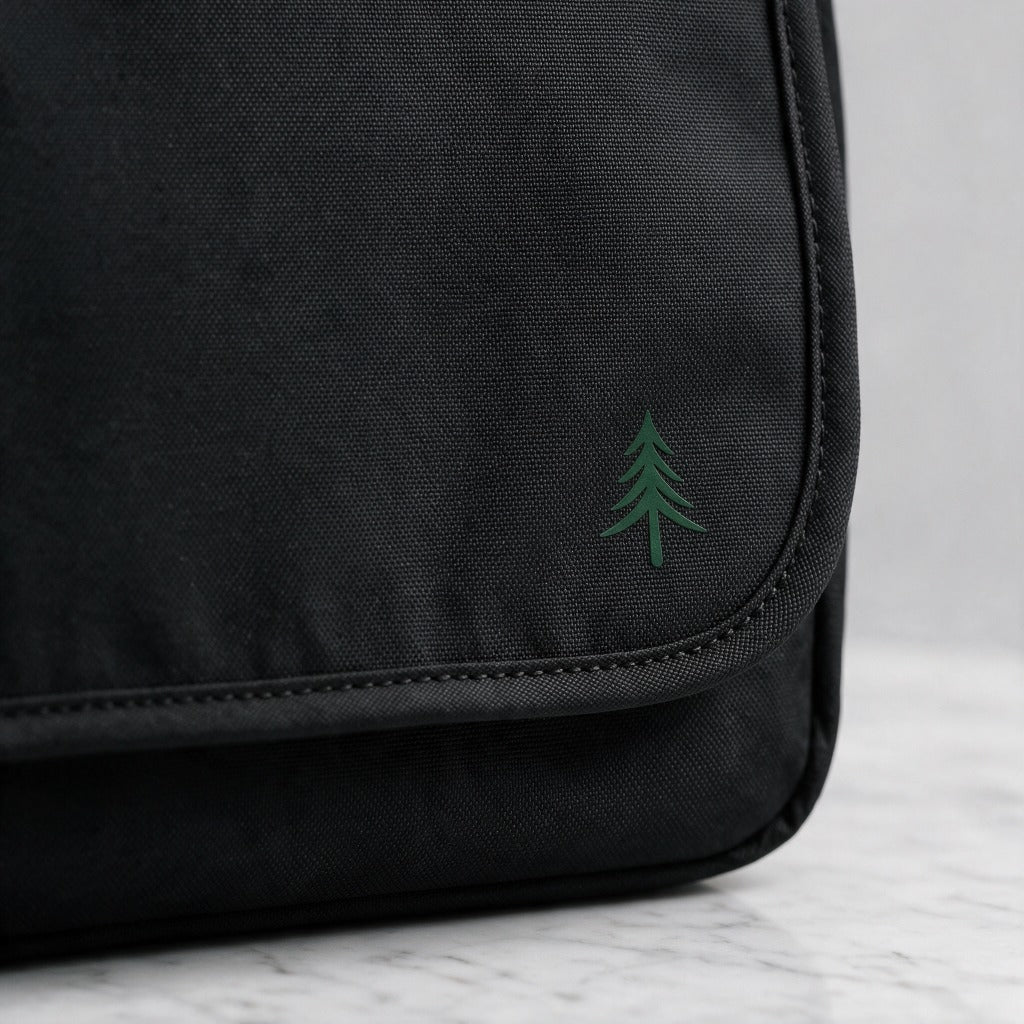 Nahaufnahme der Taschenklappe einer schwarzen FIRONA Messenger Bag aus robustem Textilgewebe mit grünem Tannenbaum-Logo und sauberer Einfassung auf einer hellen Marmorfläche.