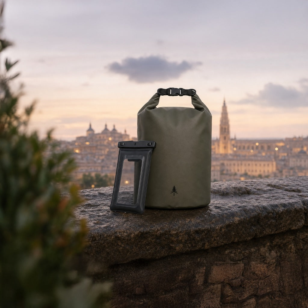 Premium-Produktfoto in der Abenddämmerung: Die olivgrüne Firona Dry Bag mit Roll-Top-Verschluss steht präsent mit der schwarzen, wasserdichten Firona Phone Pouch auf einer alten Steinmauer platziert, hinter der sich die weich gezeichnete, historische Silhouette von Toledo im sanften Licht des Abendhimmels erstreckt.